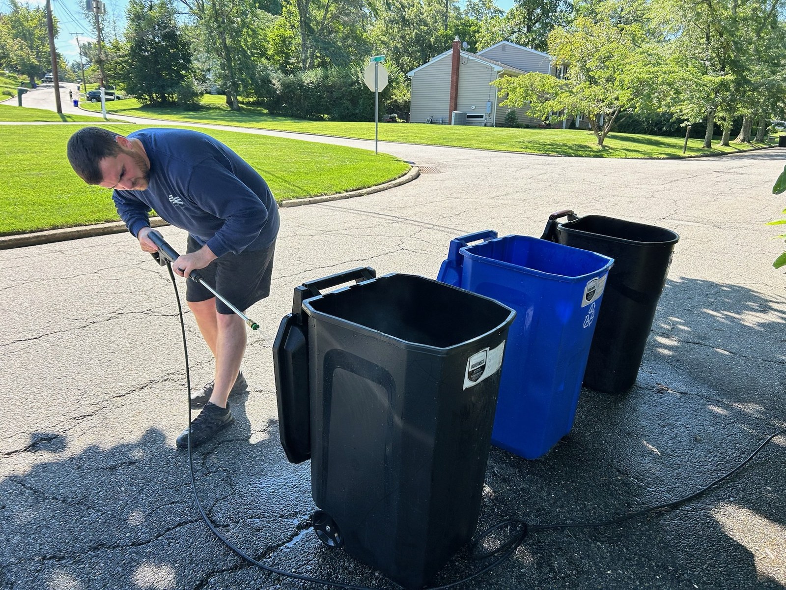 Robert power-washing four trash and recycling cans in a driveway